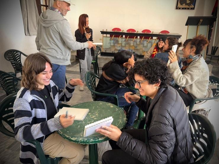 ES: Jóvenes artistas ríen y dibujan en cuadernos sobre una mesa.
FR: Jeunes artistes rient et dessinent dans des carnets sur une table.
EN: Young artists laugh and sketch in notebooks at a table.