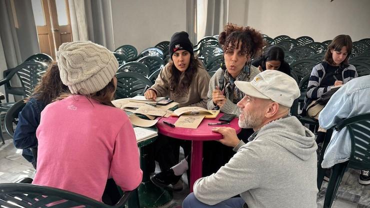 ES: Artista guía a un grupo de jóvenes que revisan bocetos.
FR: Un artiste guide un groupe de jeunes examinant des croquis.
EN: An artist guides a group of young people reviewing sketches.